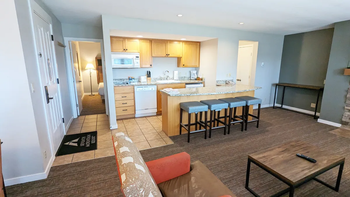 This image captures a wide view of the open-plan living space, prominently featuring the kitchen island with bar stools, the living room sofa, and the fireplace. The arrangement highlights the seamless flow between cooking, dining, and relaxing areas, all within a cozy, inviting atmosphere. The presence of countertop appliances and subtle decor details reinforce the sense of casual gathering and comfort.