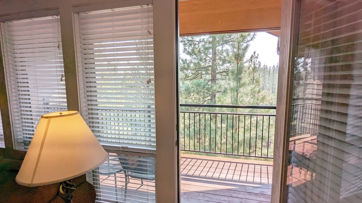 The image shows an inviting interior scene with a lamp and sliding door opening onto a deck. Through the door, trees are visible, seamlessly connecting indoor comfort with the natural outdoor environment.