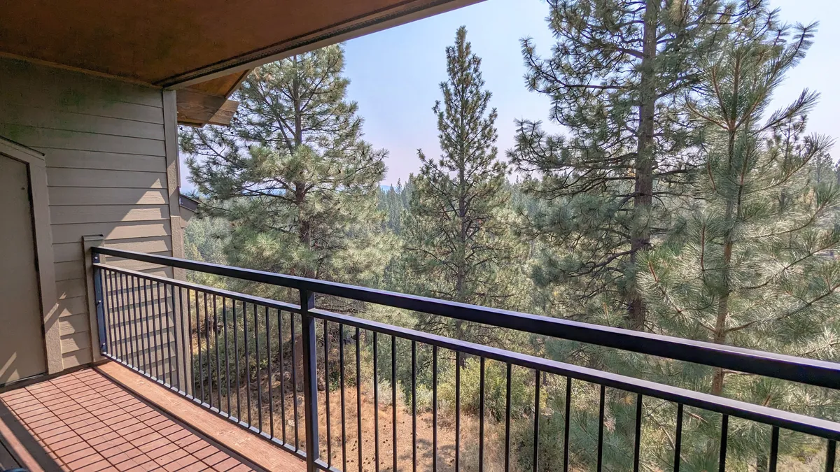 A peaceful view from the balcony shows the railing set against a rich backdrop of tall trees and open sky. The space feels secluded and enveloped by nature, making it inviting for birdwatching, morning coffee, or quiet moments of contemplation.