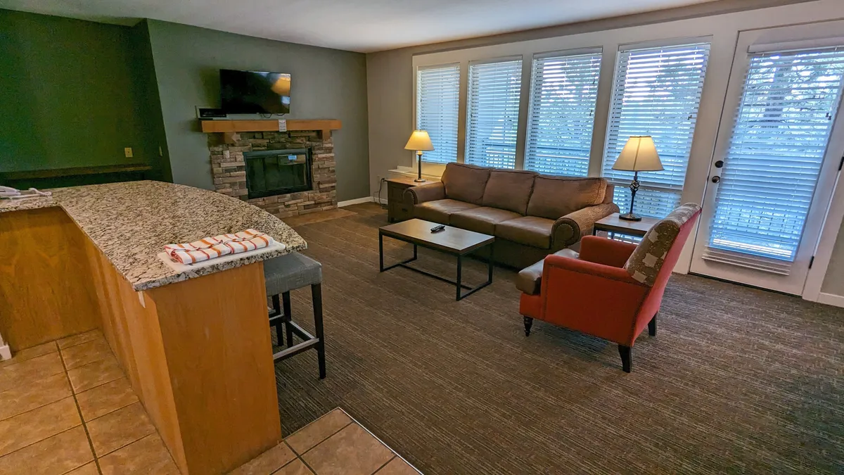 Spacious living area with a fireplace, sofa, coffee table, armchair, and expansive windows. The large windows bring in ample daylight and offer a glimpse of the natural setting, reinforcing the allure of a private balcony and forest views.